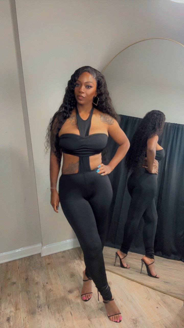 A woman modeling a black two-piece outfit consisting of a halter crop top with side cutouts and matching high-waisted leggings.